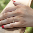 A close-up of a person's hand resting on a wooden railing outdoors, wearing a delicate gold band ring set with small round diamonds. The hand has red nail polish, and the background is a softly blurred natural setting.