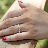 A close-up of a person's hand resting on a wooden railing outdoors, wearing a delicate gold band ring set with small round diamonds. The hand has red nail polish, and the background is a softly blurred natural setting.