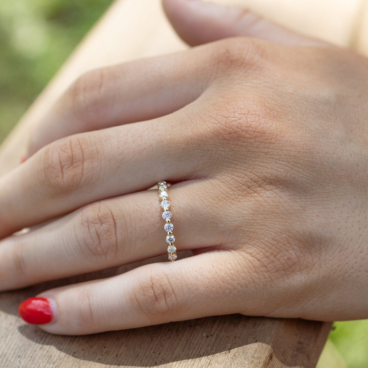A close-up of a person's hand resting on a wooden railing outdoors, wearing a delicate gold band ring set with small round diamonds. The hand has red nail polish, and the background is a softly blurred natural setting.