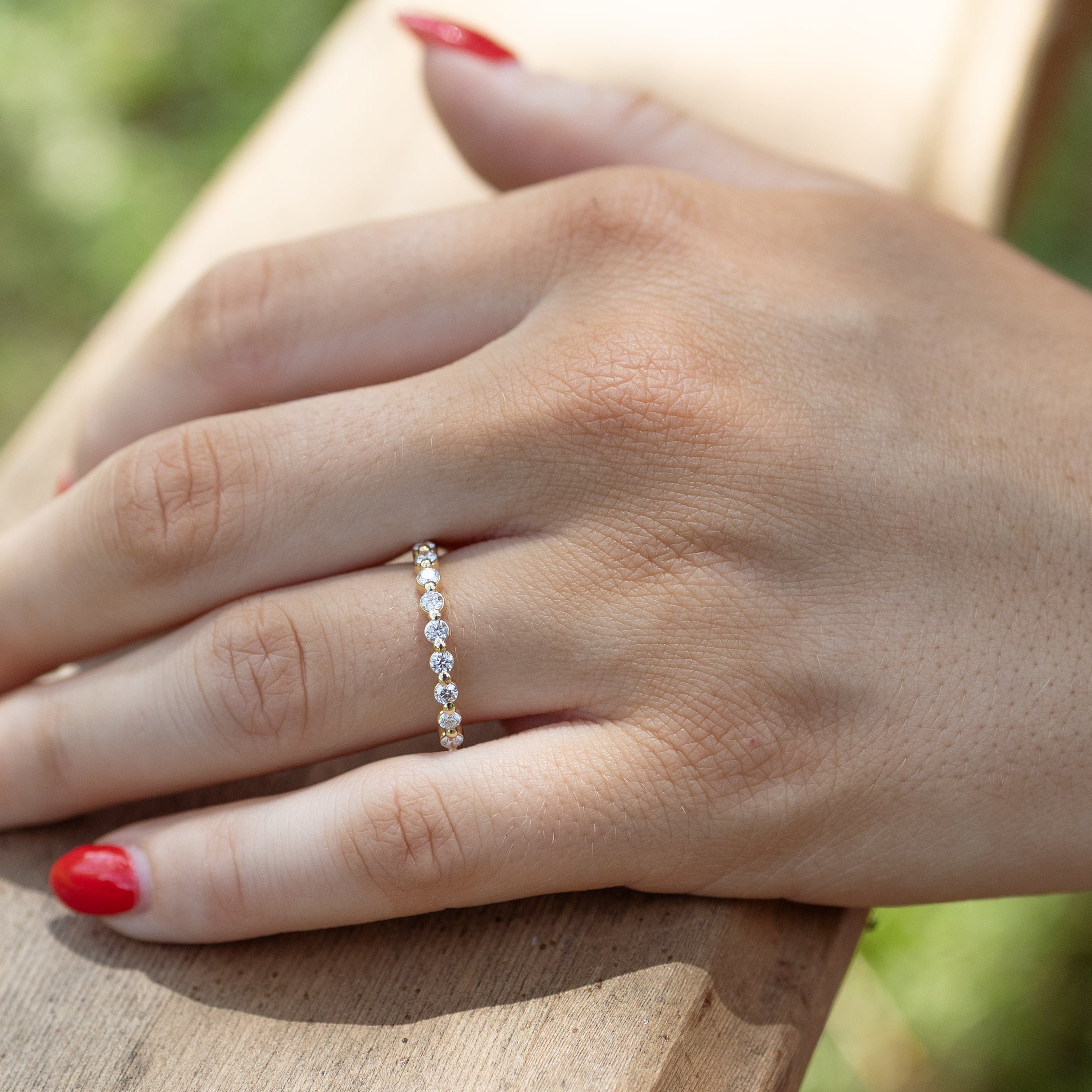 A close-up of a person's hand resting on a wooden railing outdoors, wearing a delicate gold band ring set with small round diamonds. The hand has red nail polish, and the background is a softly blurred natural setting.