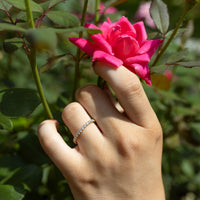 A close-up of a hand wearing a delicate diamond band ring, gently touching a vibrant pink rose in a garden with green foliage.