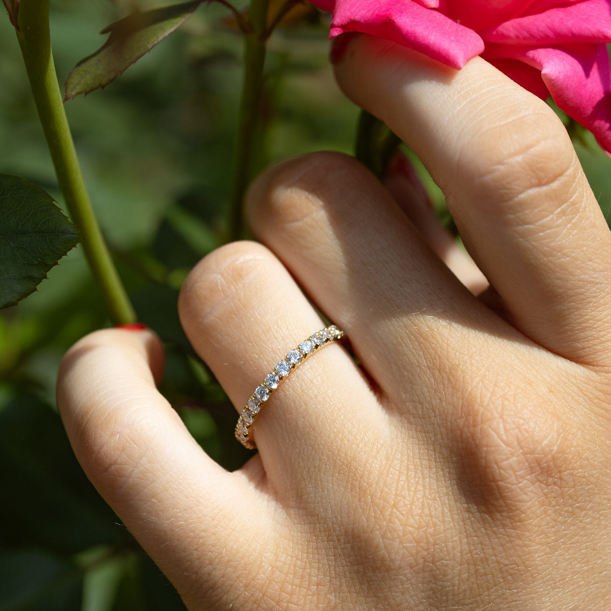 A close-up of a hand wearing a delicate diamond band ring, gently touching a vibrant pink rose in a garden with green foliage.