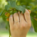 A close-up of a hand wearing a delicate gold band ring with small round diamonds, set against a soft-focus background of green leaves and small yellow-orange berries.