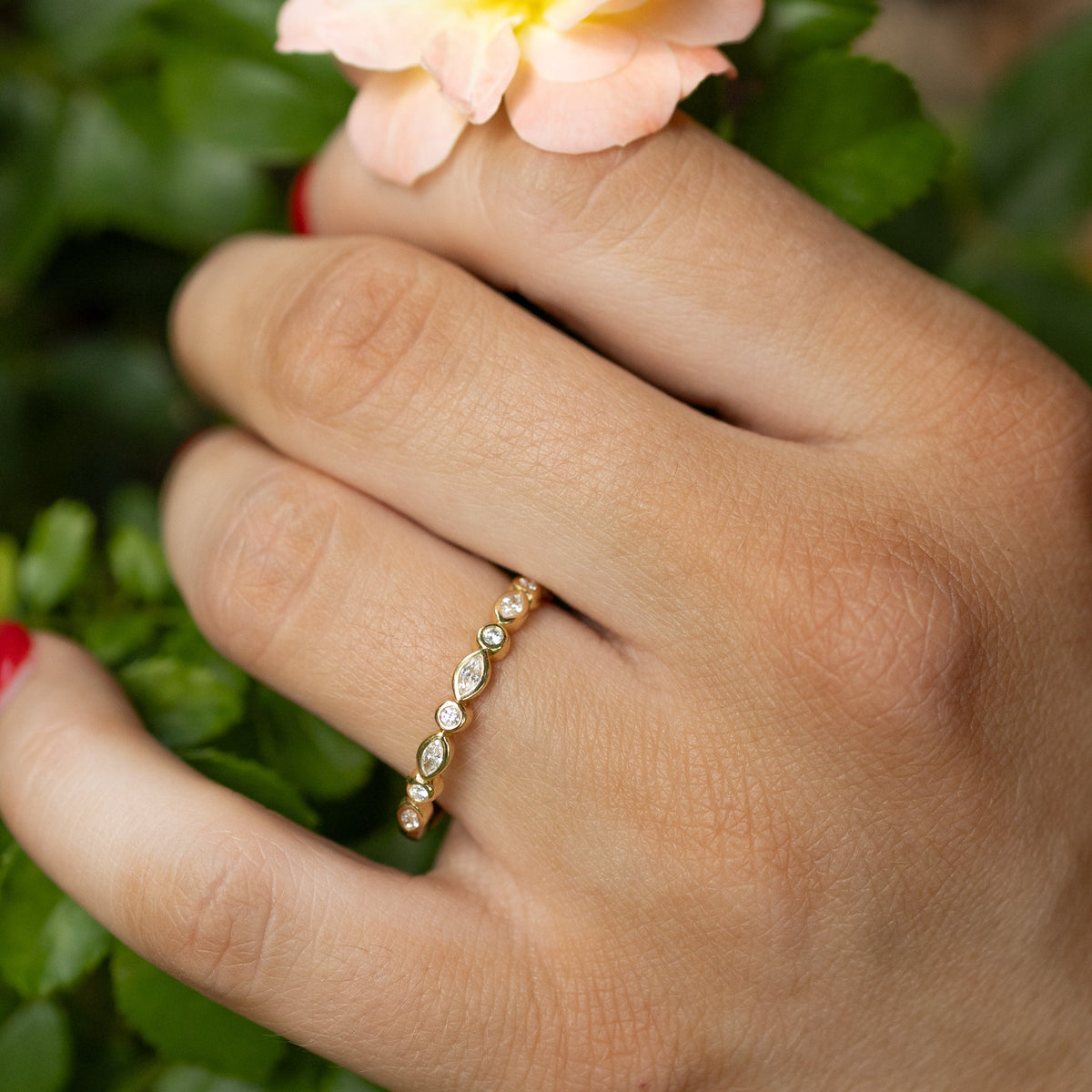 A close-up of a hand wearing a delicate gold ring with small marquise and round diamonds, resting near a pale peach rose among green leaves.