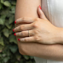 A close-up of a person's hand resting on their elbow, wearing a delicate gold band ring with small diamonds. The person has red-painted fingernails and is wearing a white sleeveless top, with a soft-focus background of green ivy.