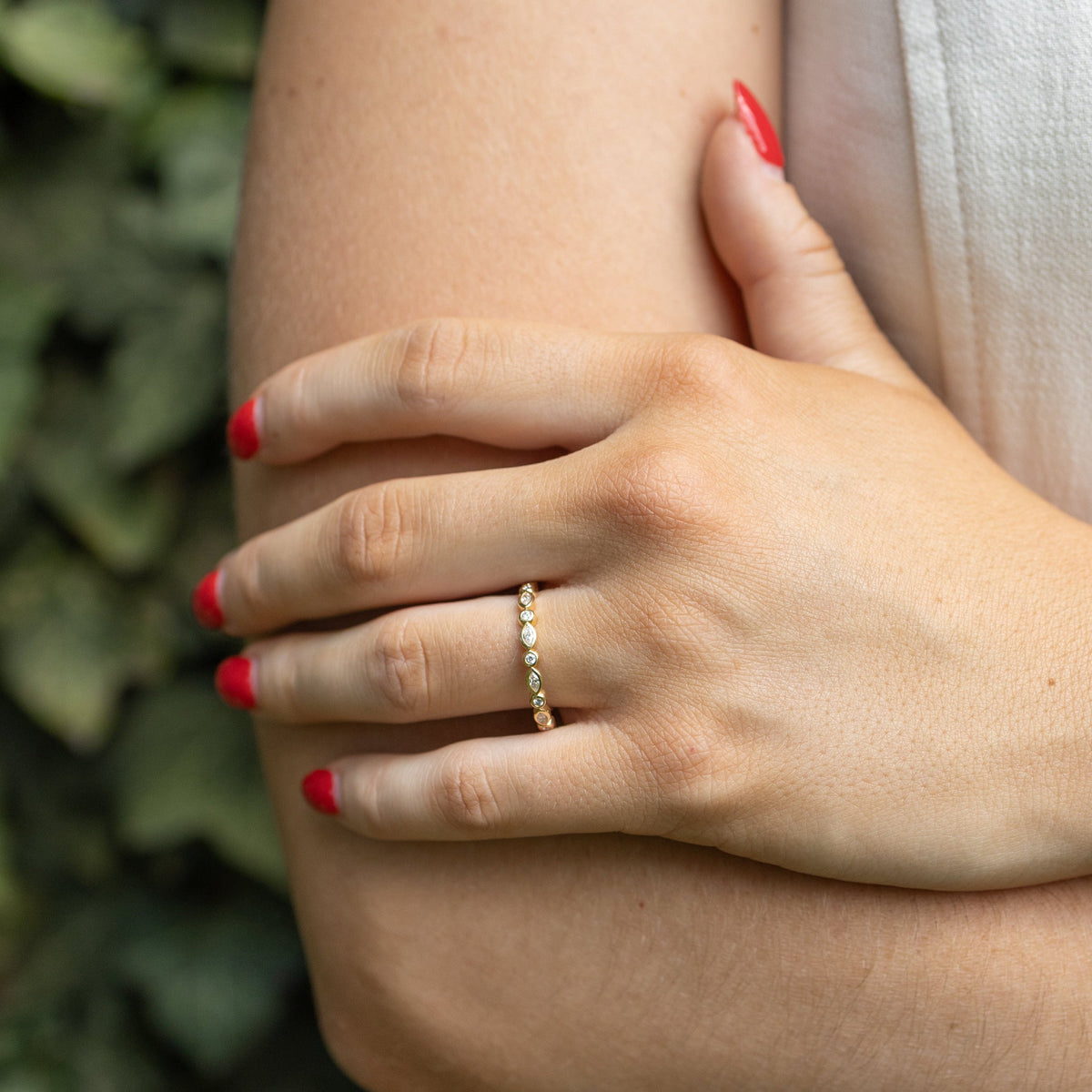 A close-up of a person's hand resting on their elbow, wearing a delicate gold band ring with small diamonds. The person has red-painted fingernails and is wearing a white sleeveless top, with a soft-focus background of green ivy.