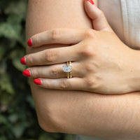 A close-up lifestyle shot of a person's hand resting on their arm, wearing a gold engagement ring with a elongated cushion diamond. The background is a soft-focus green ivy wall, and the person is wearing a white sleeveless top.