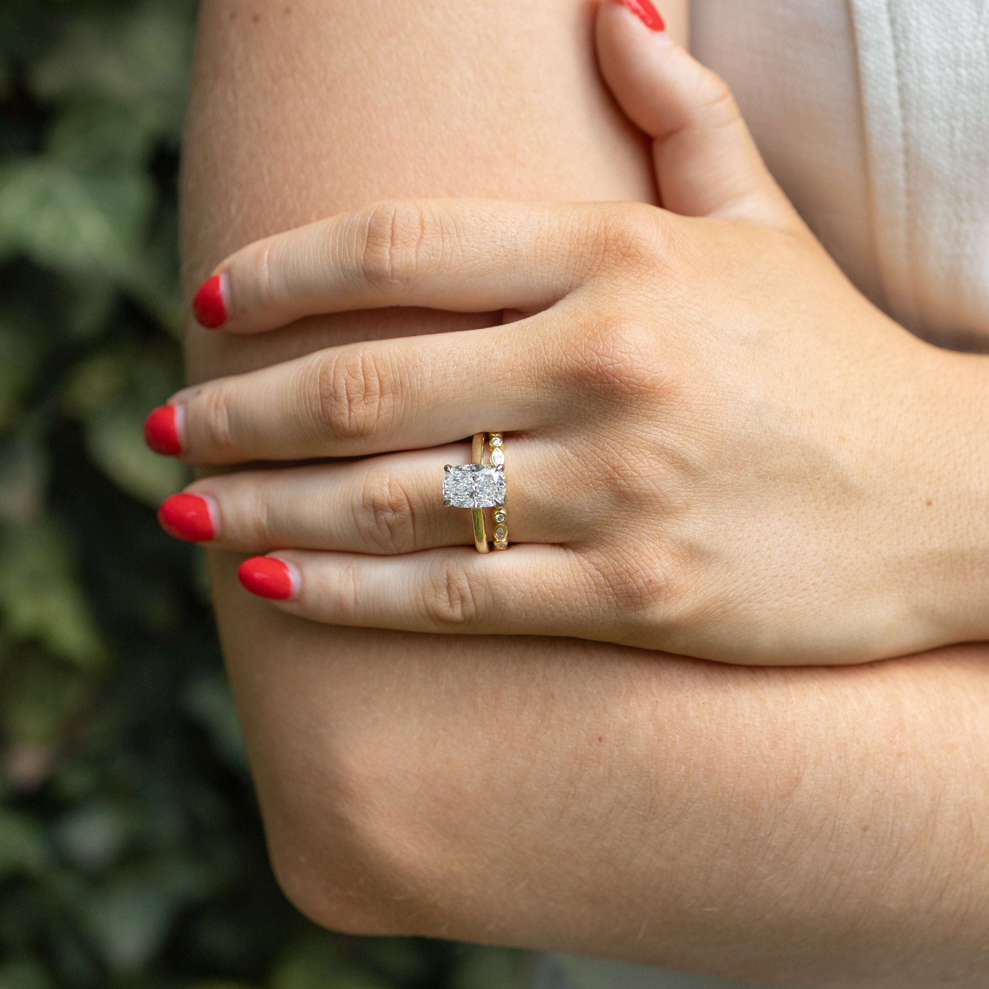 A close-up lifestyle shot of a person's hand resting on their arm, wearing a gold engagement ring with a elongated cushion diamond. The background is a soft-focus green ivy wall, and the person is wearing a white sleeveless top.