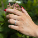 A close-up of a person's hand with red nail polish wearing a thin white gold diamond ring set with small round diamonds, posed against a soft-focus background of green leaves and pink hydrangea flowers.