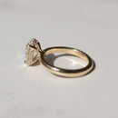 side view of a yellow gold engagement ring with a hidden halo and oval center diamond on a white marble backdrop