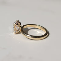 side view of a yellow gold engagement ring with a hidden halo and oval center diamond on a white marble backdrop