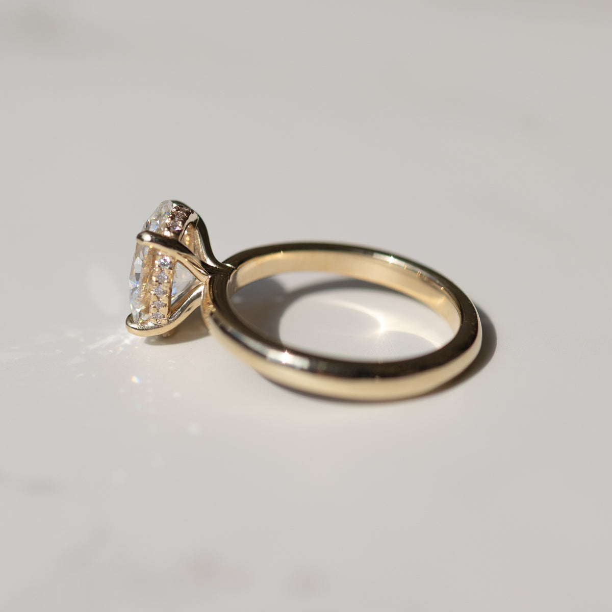 side view of a yellow gold engagement ring with a hidden halo and oval center diamond on a white marble backdrop