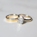 A gold engagement ring with an oval-cut diamond in a prong setting, shown next to a plain gold band on a light gray reflective surface.