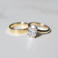 A gold engagement ring with an oval-cut diamond in a prong setting, shown next to a plain gold band on a light gray reflective surface.