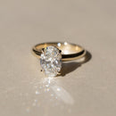 An oval-cut diamond engagement ring set in a yellow gold band, resting on a neutral beige surface with a soft reflection and shadow.
