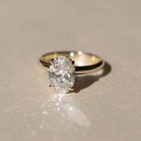 An oval-cut diamond engagement ring set in a yellow gold band, resting on a neutral beige surface with a soft reflection and shadow.