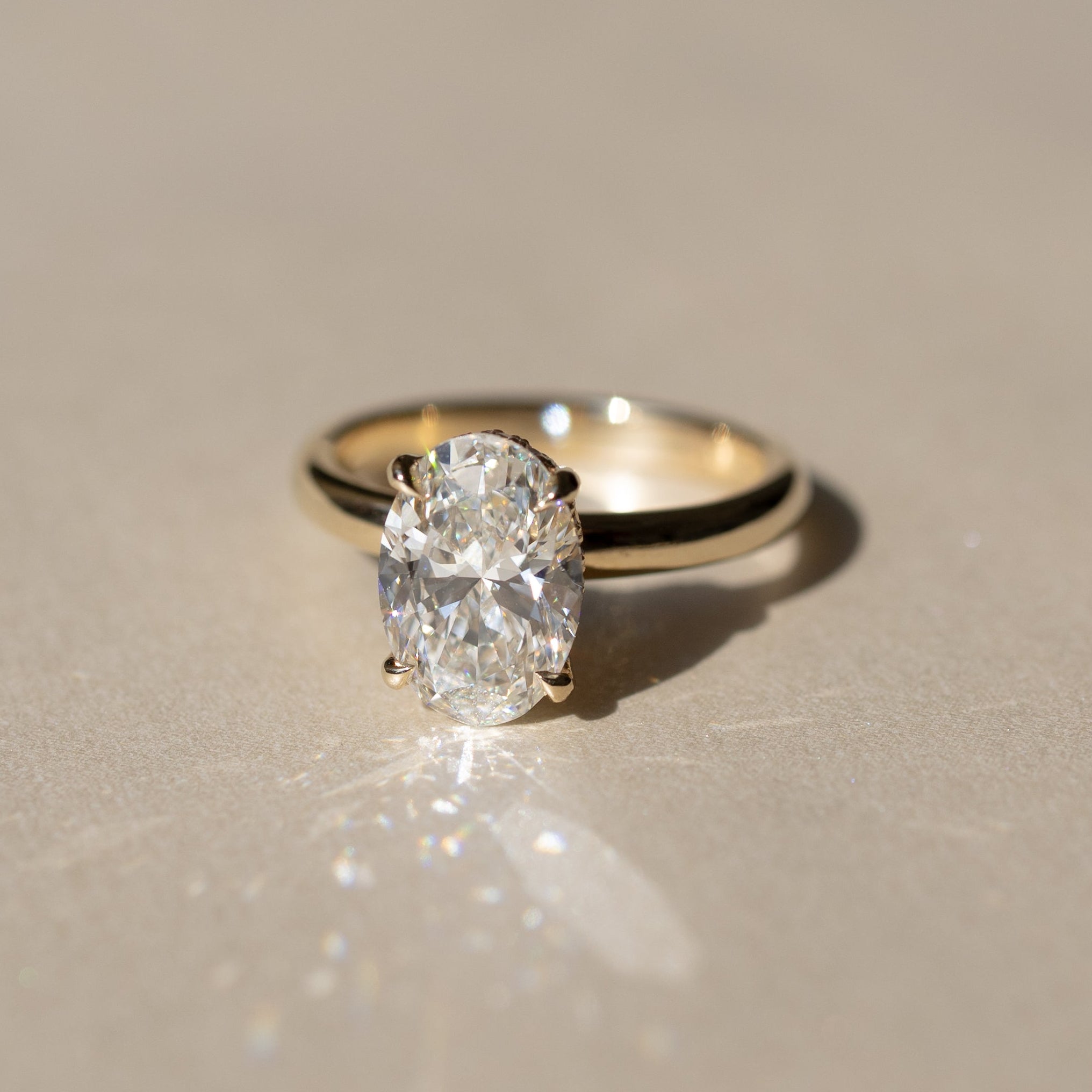 An oval-cut diamond engagement ring set in a yellow gold band, resting on a neutral beige surface with a soft reflection and shadow.