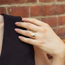 A close-up of a woman's hand wearing a gold ring with a large oval diamond, resting on her shoulder against a red brick wall background.