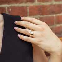 A close-up of a woman's hand wearing a gold ring with a large oval diamond, resting on her shoulder against a red brick wall background.