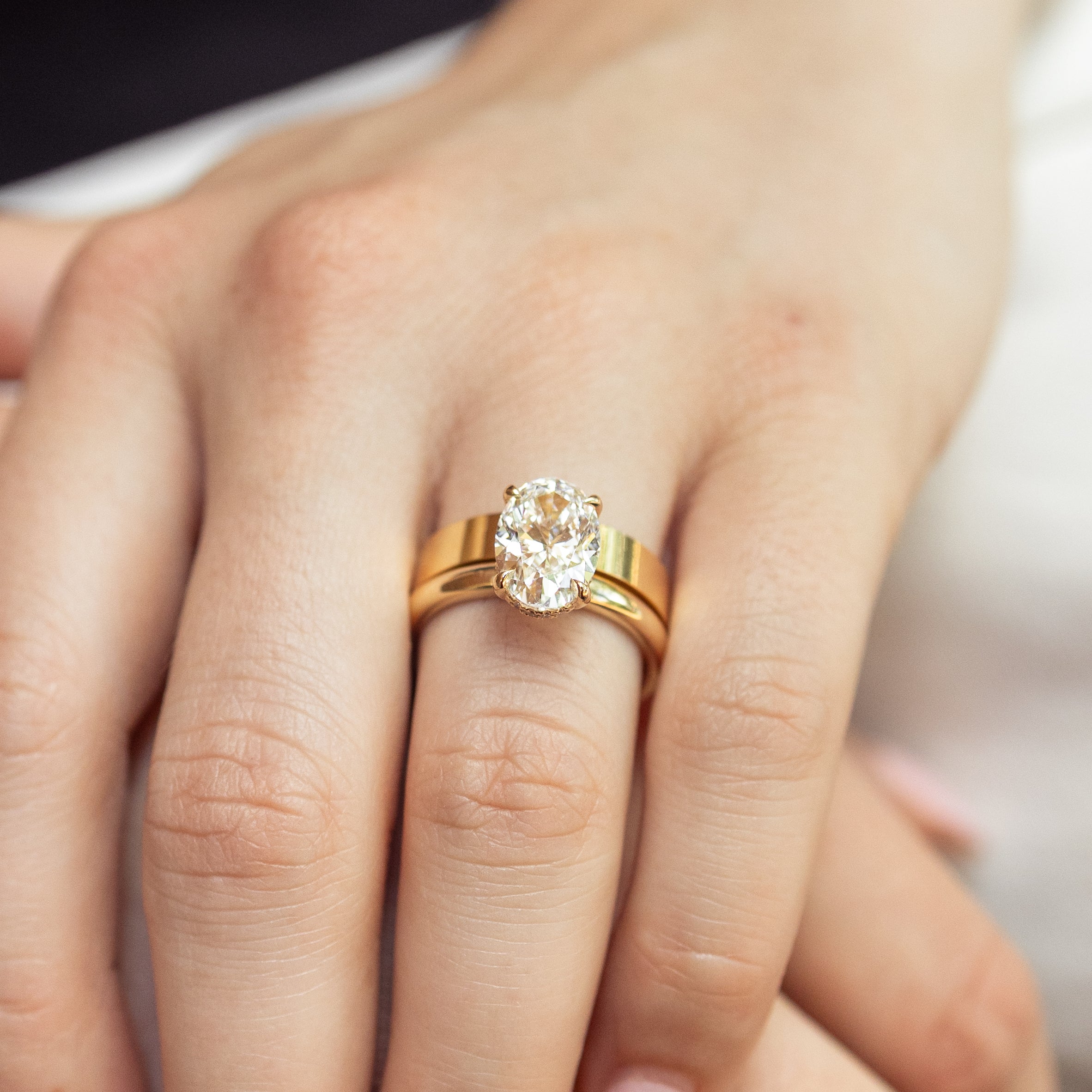 A close-up of a person's hand wearing a yellow gold ring set with an oval-cut diamond, with a matching gold flat band underneath.