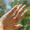 A close-up of a hand wearing a gold engagement ring with a oval-cut diamond, set against a blurred outdoor background of green foliage and blue sky.