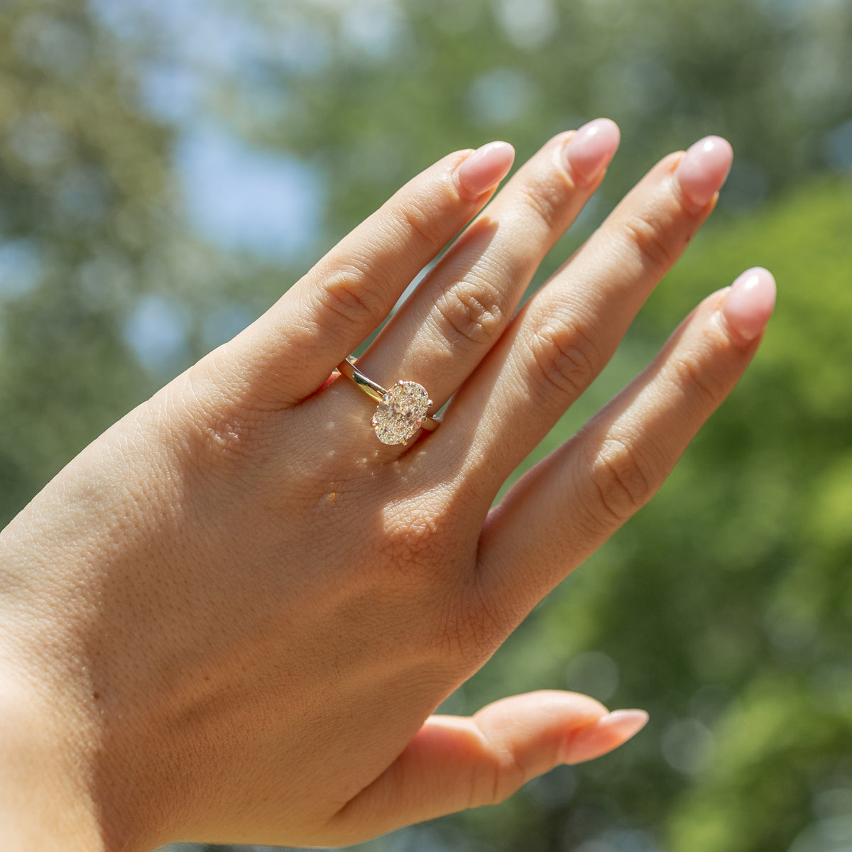 A close-up of a hand wearing a gold engagement ring with a oval-cut diamond, set against a blurred outdoor background of green foliage and blue sky.